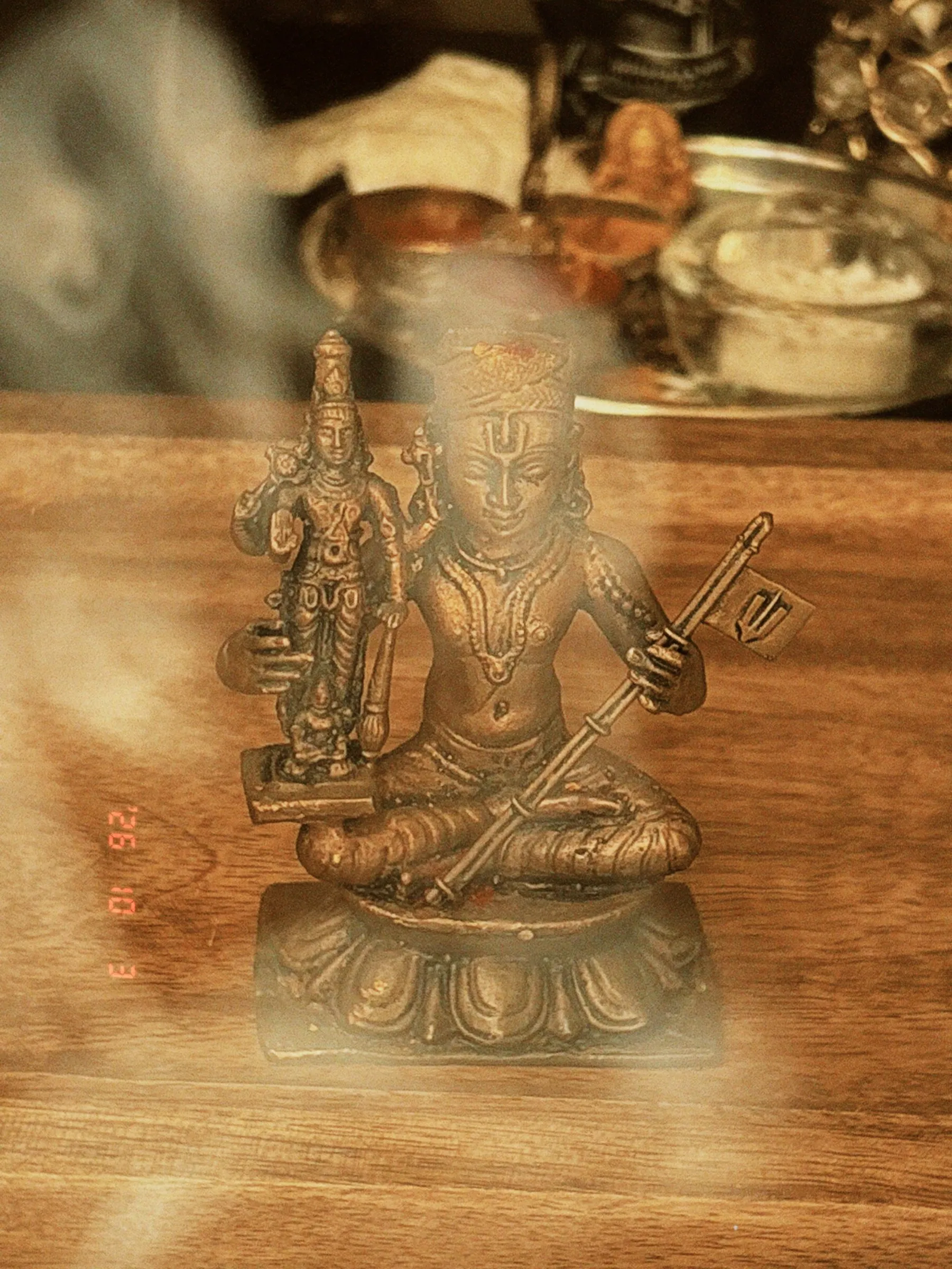 A small bronze deity statue on a wooden altar, softened by drifting incense smoke