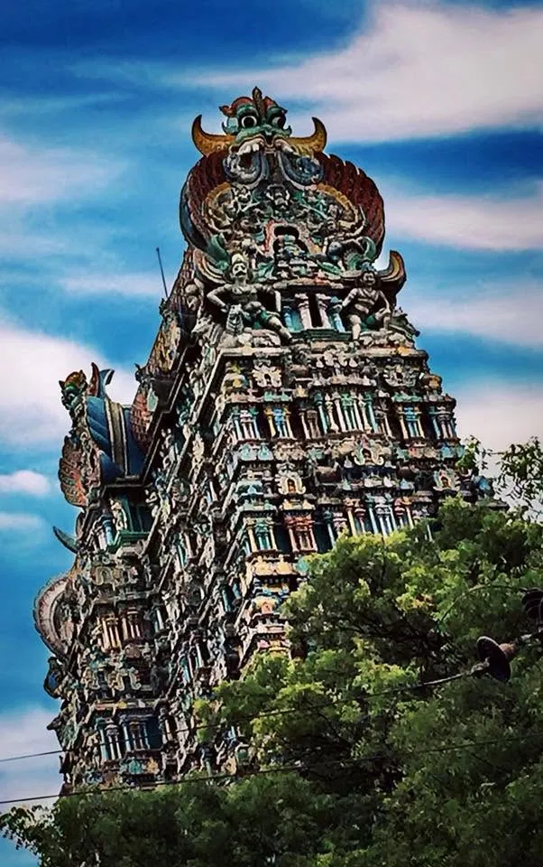 A tall, intricately carved temple tower rising above trees against a vivid blue sky with clouds