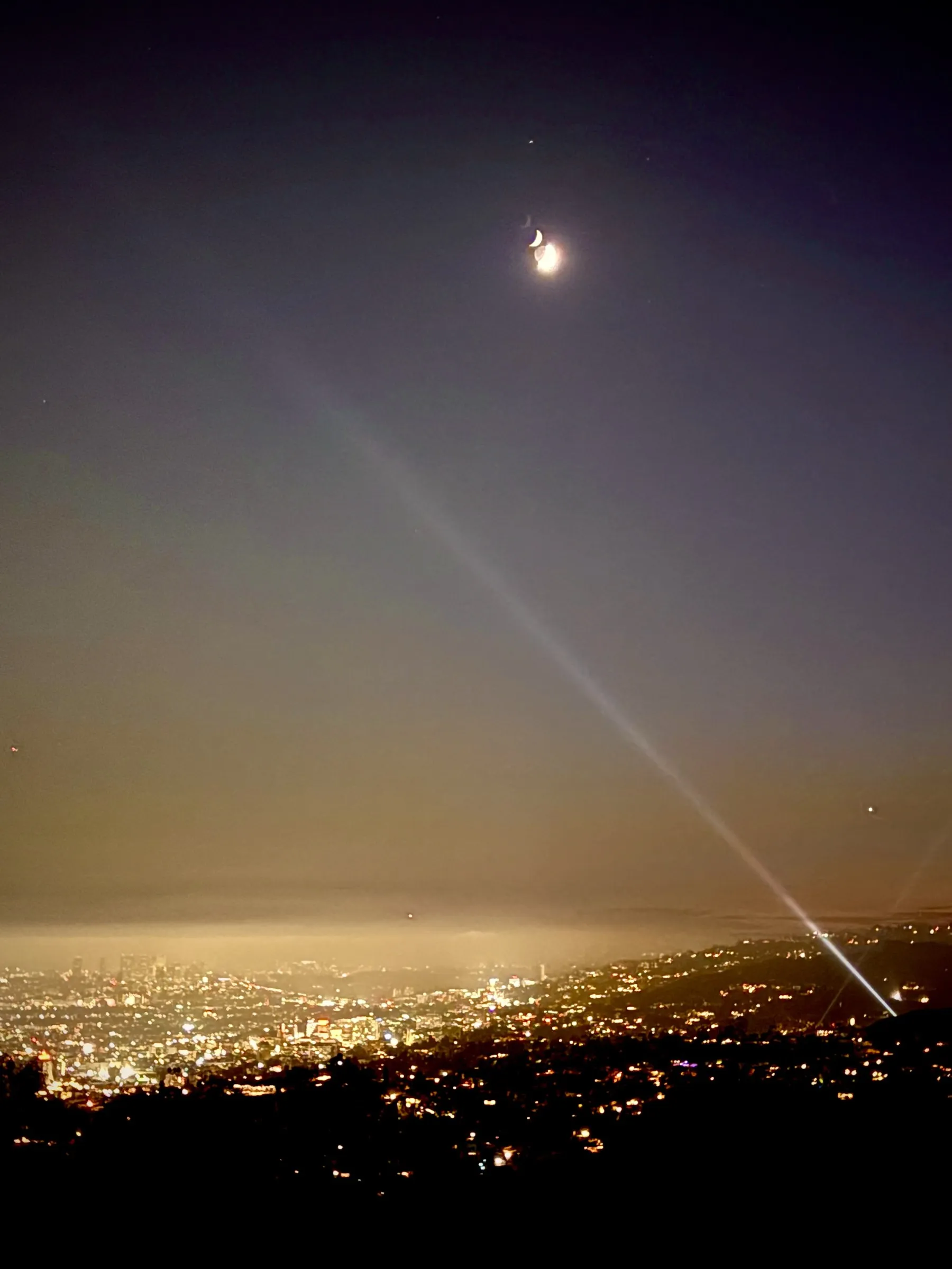 A crescent moon over the lights of Los Angeles at night, with a searchlight beam across the sky