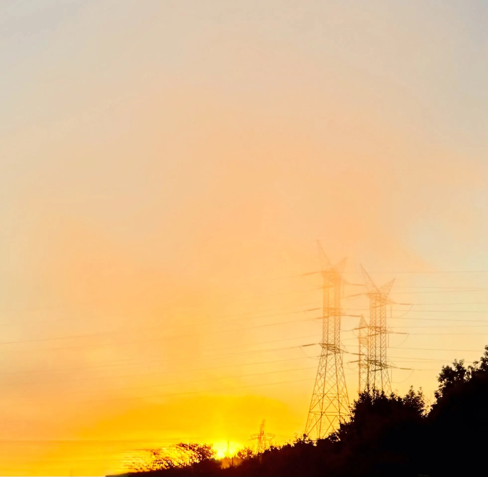 Power-line pylons and a dark tree line silhouetted against a hazy orange and gold sunset