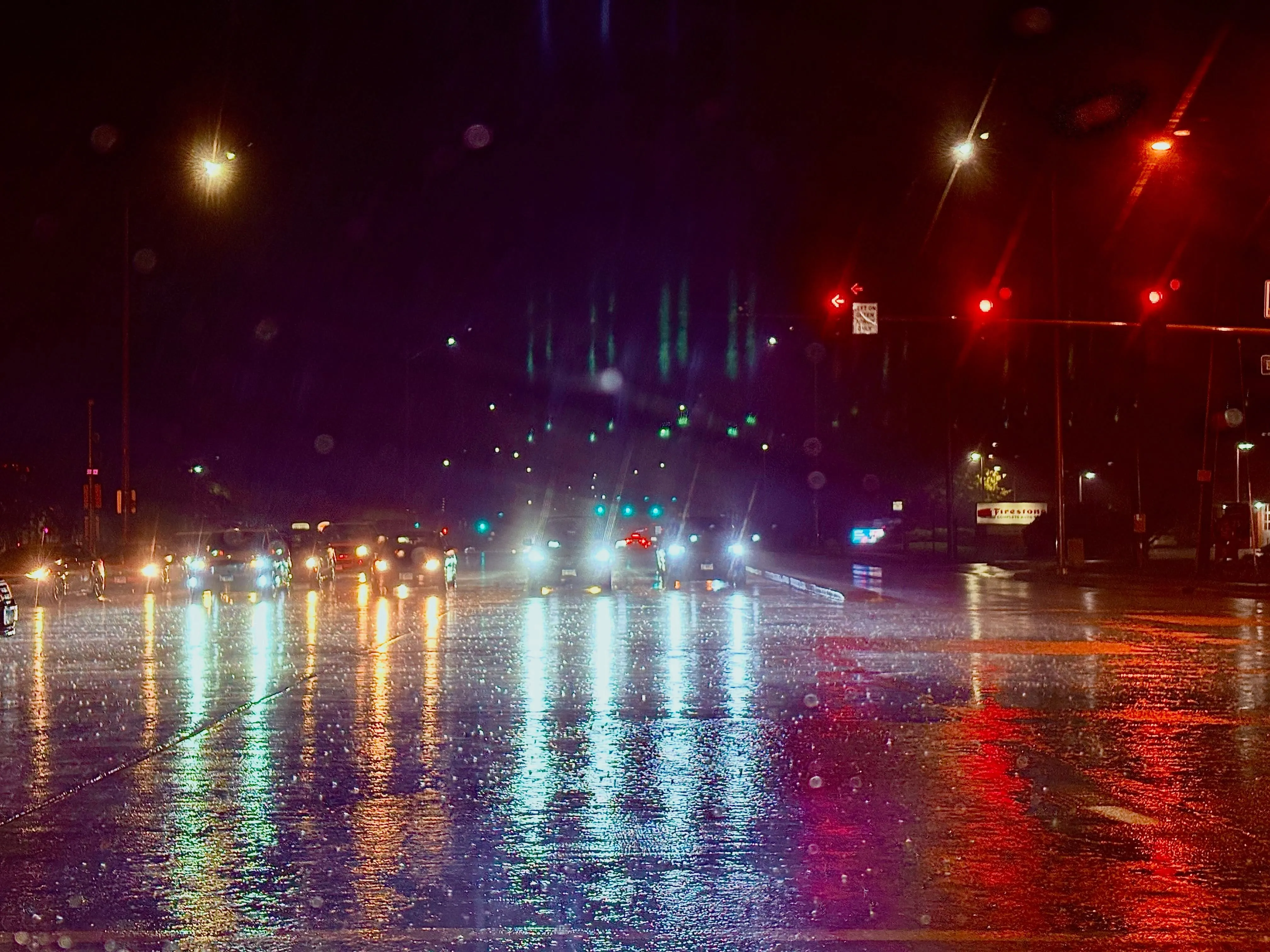 A rain-soaked intersection at night, headlights and red signals stretched into long reflections on the wet road