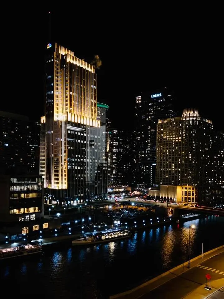 Downtown Chicago towers glowing at night above the dark river and its lit walkways