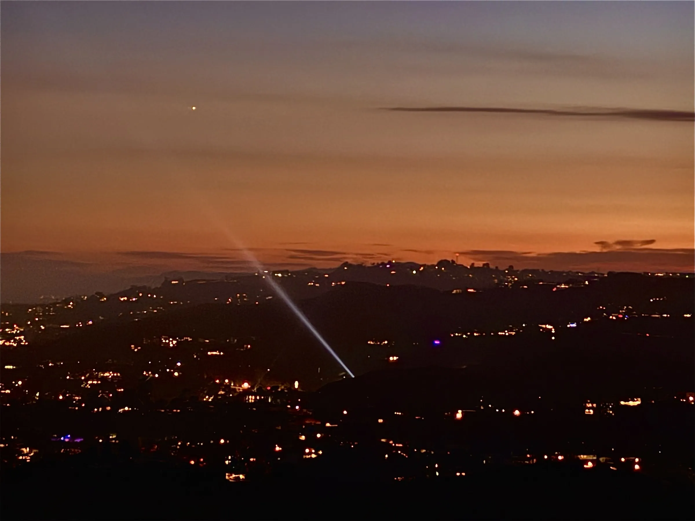 A searchlight beam cutting across an orange dusk sky above the glittering lights of Los Angeles