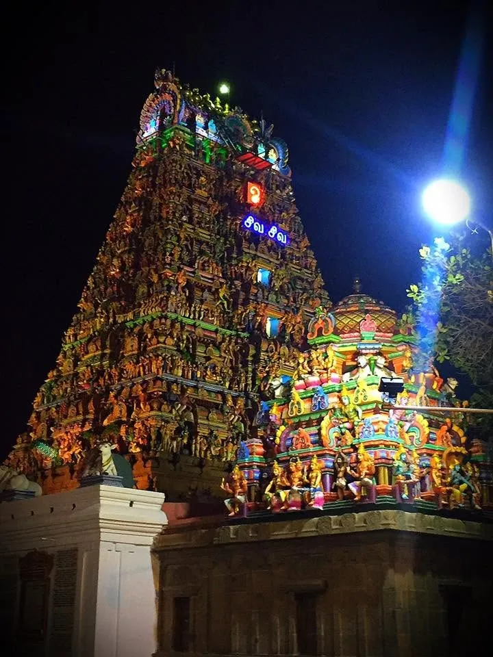 A temple tower covered in colorful sculptures, lit for a festival at night with a bright lamp beside it