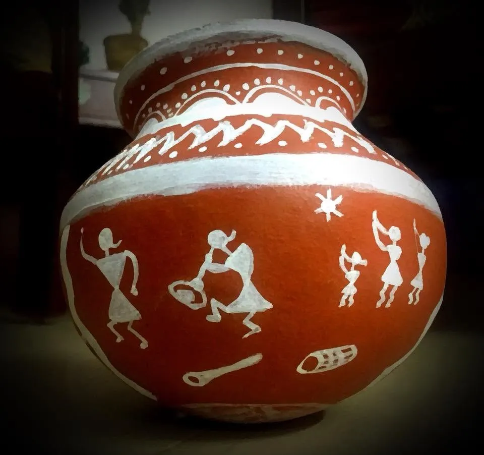 A terracotta pot painted with white Warli folk-art figures dancing and playing music